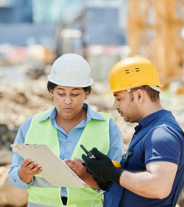 Two construction workers discussing a project on-site, wearing safety gear including hard hats and reflective vests.